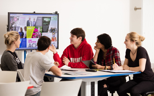 Group of five students (two males, three females) sat around a white desk studying a television screen