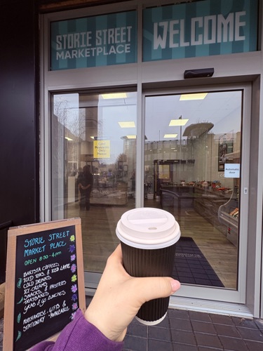 Student holding a takeaway cup of coffee at the entrance to the Storie Street Marketplace coffee shop